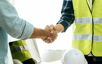 A facilities manager and a contractor wearing a high vis vest shake hands over a laptop