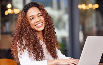 A young female-presenting facilities manager with curly hair smiles at the camera while working on a laptop