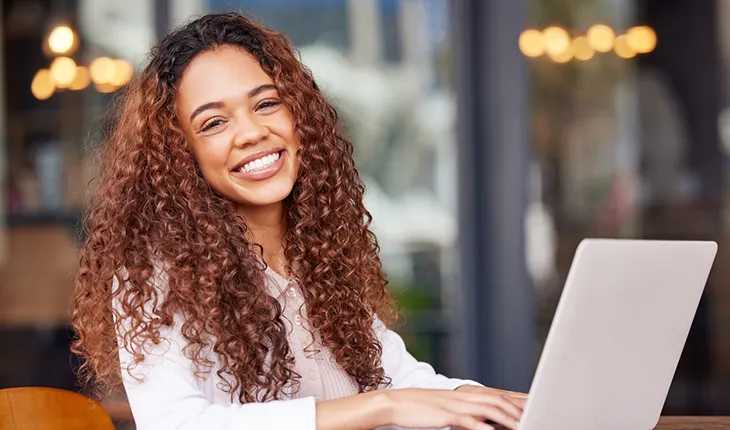 A young female-presenting facilities manager with curly hair smiles at the camera while working on a laptop