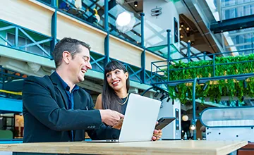 A man and a woman, both wearing suits, look at data on a laptop while operating in a building adorned with hanging plants
