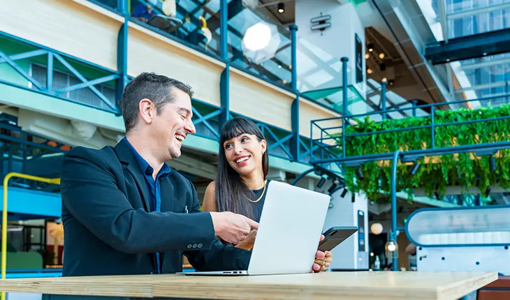A man and a woman, both wearing suits, look at data on a laptop while operating in a building adorned with hanging plants