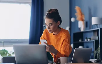 A woman in an orange jumper holds a mobile phone while looking at a laptop screen