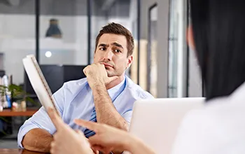 A suspicious facility manager at a desk raises an eyebrow at a potential software partner, obscured, who is showing a contract