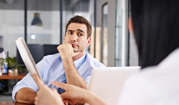 A suspicious facility manager at a desk raises an eyebrow at a potential software partner, obscured, who is showing a contract