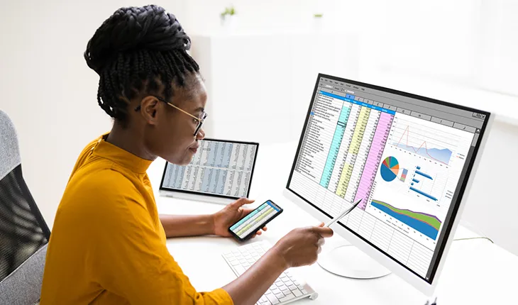 A young woman compares data on a computer with data on a phone screen