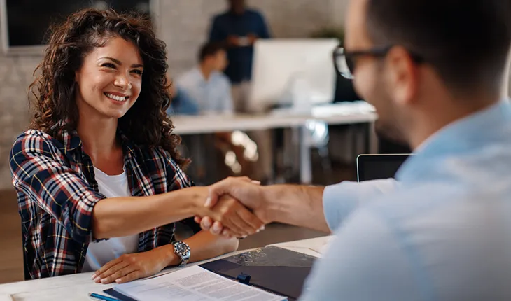 A young woman in a t shirt and flannel shirt shakes the hand of a man wearing glasses at the end of a successful job interview