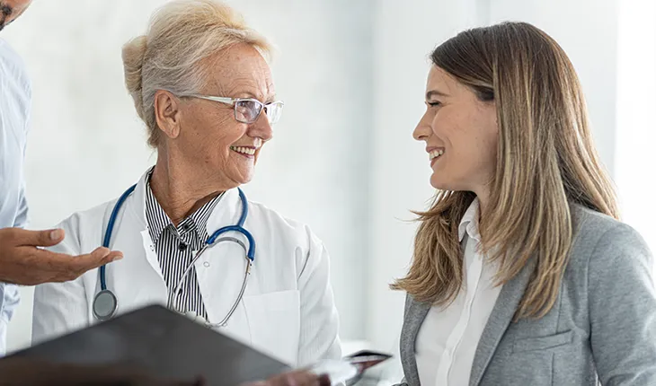 A doctor wearing a white coat and with a stethescope draped over her shoulders speaks to a facilites manager wearing a grey blazer