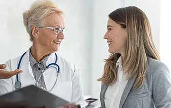 A doctor wearing a white coat and with a stethescope draped over her shoulders speaks to a facilites manager wearing a grey blazer