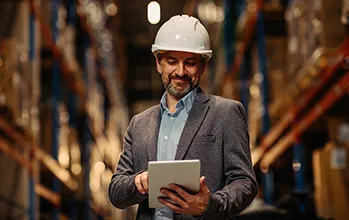A facilities manager wearing a suit and a hard hat reviews asset data on an ipad and smiles to themselves