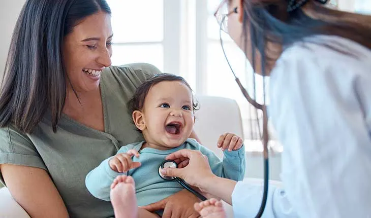 A baby smiles, held by its mother as a doctor listens to its heartbeat on a stethoscope