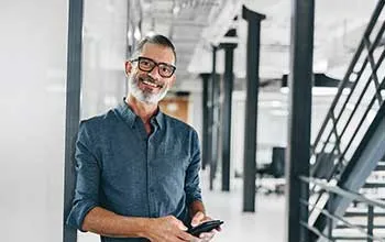 A facilities manager holding a phone smiles at the camera while leaning against a wall in a modern office building