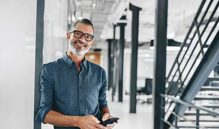 A facilities manager holding a phone smiles at the camera while leaning against a wall in a modern office building