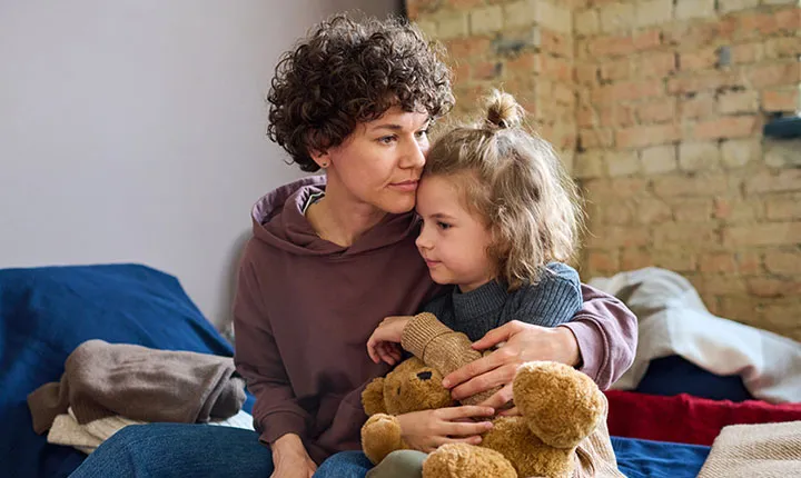 A woman in a shelter holds her young child who is gripping a teddy bear.