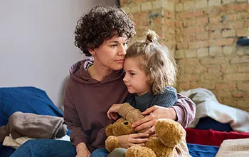 A woman in a shelter holds her young child who is gripping a teddy bear.