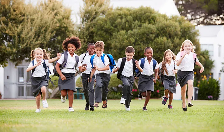 A group of primary school children run across a green oval towards the camera