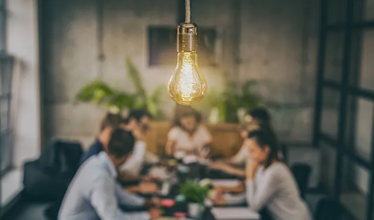 a business meeting is observed from above, with a lit lightbulb in the foreground.