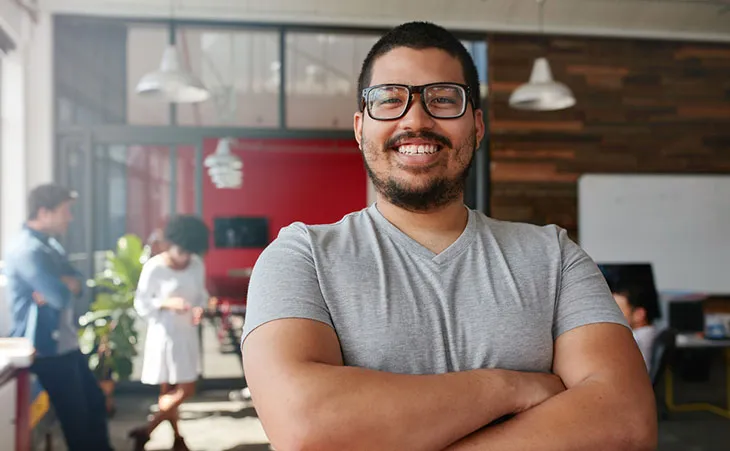 A facilities manager wearing a grey t shirt and glasses smiles at the camera with crossed arms