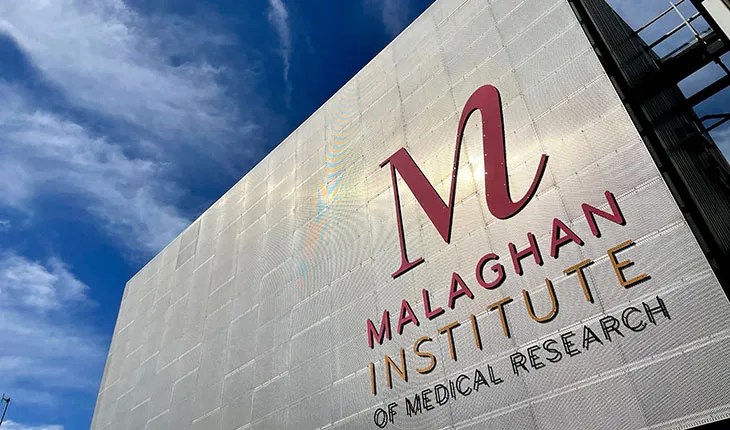 A scientist in a lab coat looks into a microscope, wearing safety glasses and safety gloves and holding a sample of something in a vial