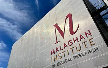 A scientist in a lab coat looks into a microscope, wearing safety glasses and safety gloves and holding a sample of something in a vial