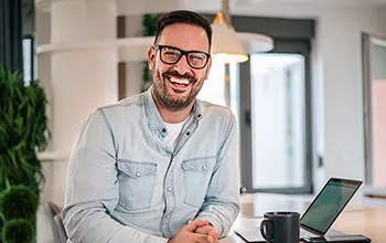 A facilities manager smiles at the camera from a desk with a laptop and a mug