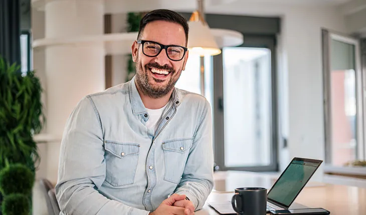 A facilities manager smiles at the camera from a desk with a laptop and a mug