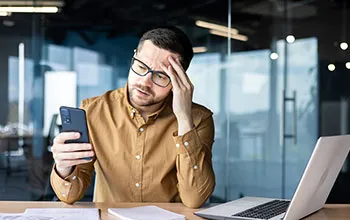 A facilities manager has a stressed look as he looks at his mobile phone, papers strewn across the desk, and a laptop open.