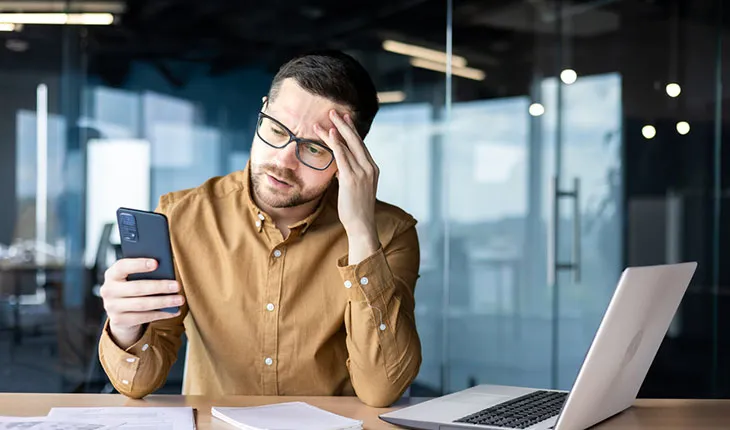 A facilities manager has a stressed look as he looks at his mobile phone, papers strewn across the desk, and a laptop open.