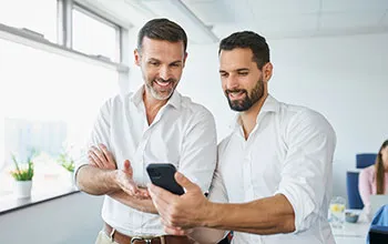 Two middle aged male facility users look at a smartphone screen and smile, one of them holding the phone and the other gesturing towards the screen