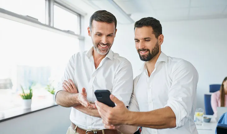 Two middle aged male facility users look at a smartphone screen and smile, one of them holding the phone and the other gesturing towards the screen