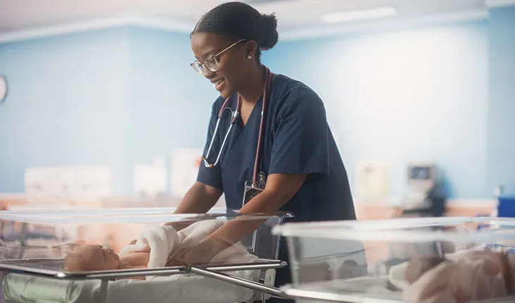 A doctor visits a maternity ward to check on an infant, both are smiling at each other and the dr wears a stethescope around her neck.