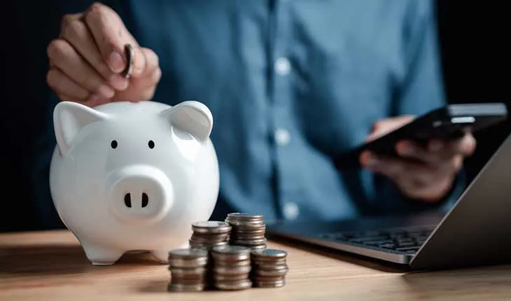 A facilities manager puts a coin into a white piggybank, while working on a calculator