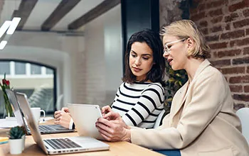Two facilities managers look at a tablet device while sitting in front of laptops, one is pointing to the tablet screen and explaining something.