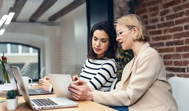 Two facilities managers look at a tablet device while sitting in front of laptops, one is pointing to the tablet screen and explaining something.