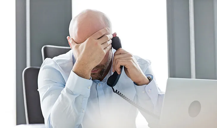 A facilities manager drops his head into his hand in frustration while talking on a landline phone