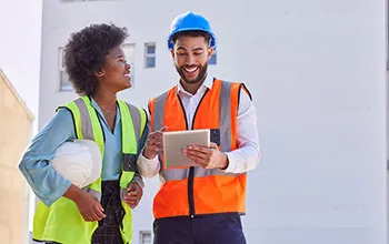 Two facility managers in high vis safety vests enjoy a lively discussion while looking at an ipad
