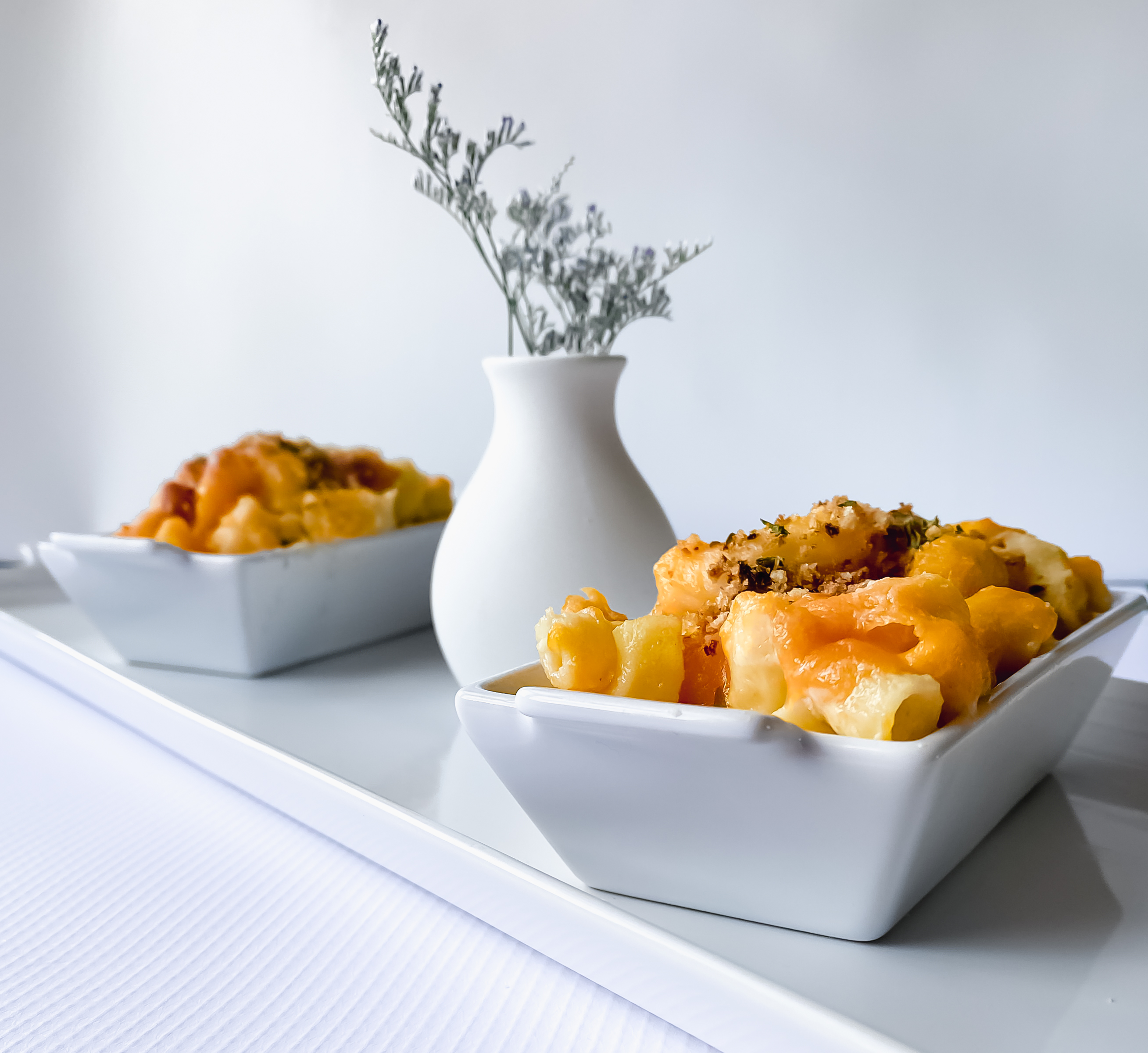 Two white dishes filled with golden Mac and cheese topped with bread crumbs. A white vase with small purple flowers is between them. The macaroni and cheese and vase are situated on a white platter with a white background and white flat lay. The image is in the style of light and bright. 