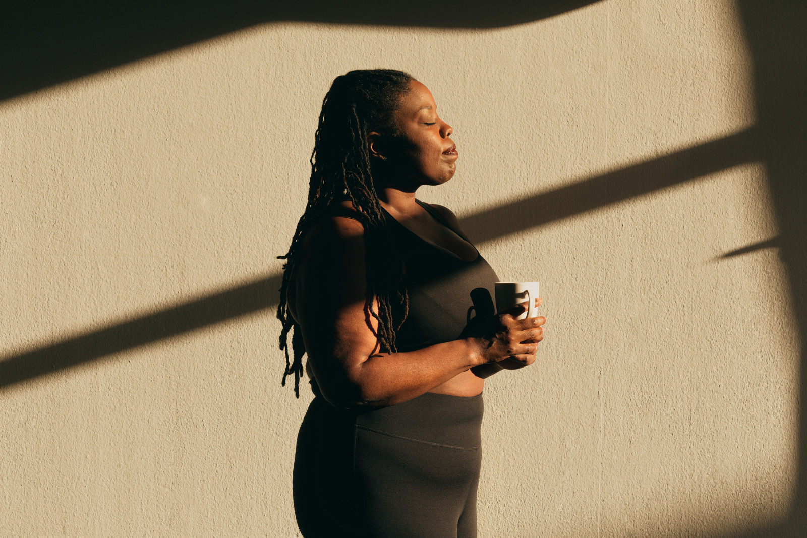 A woman holding a mug stands in sunlight with her eyes closed.