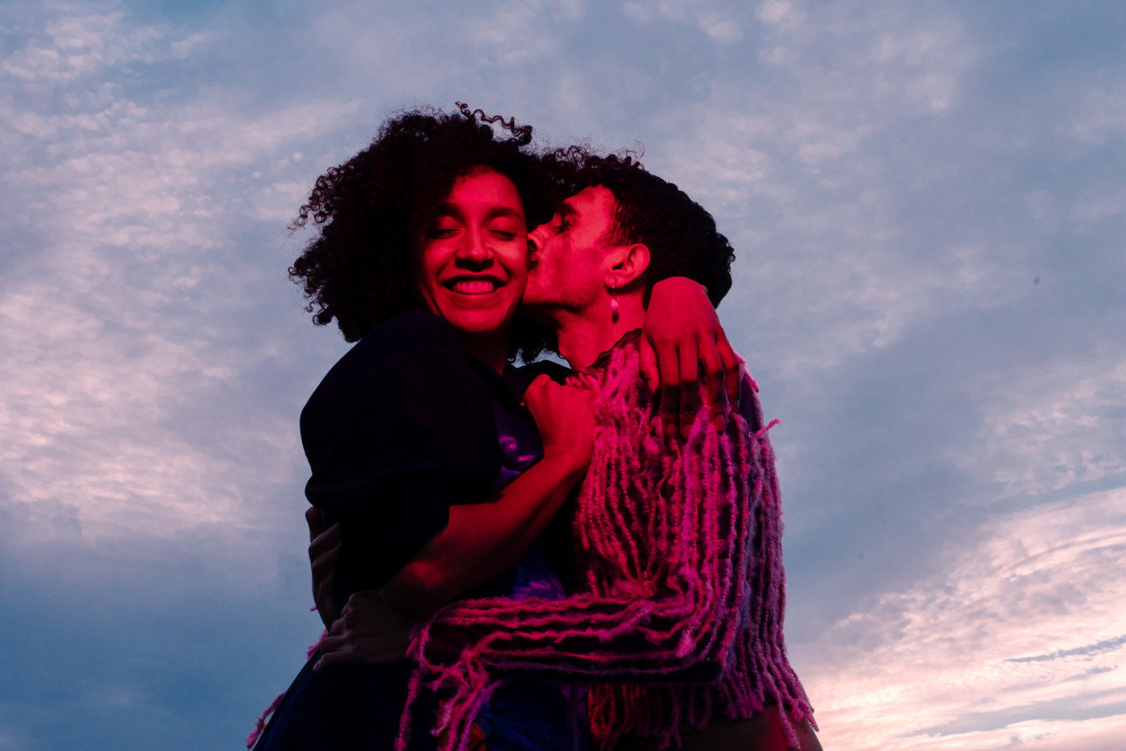 Two people embrace under colourful lights, in front of a cloudy blue sky.