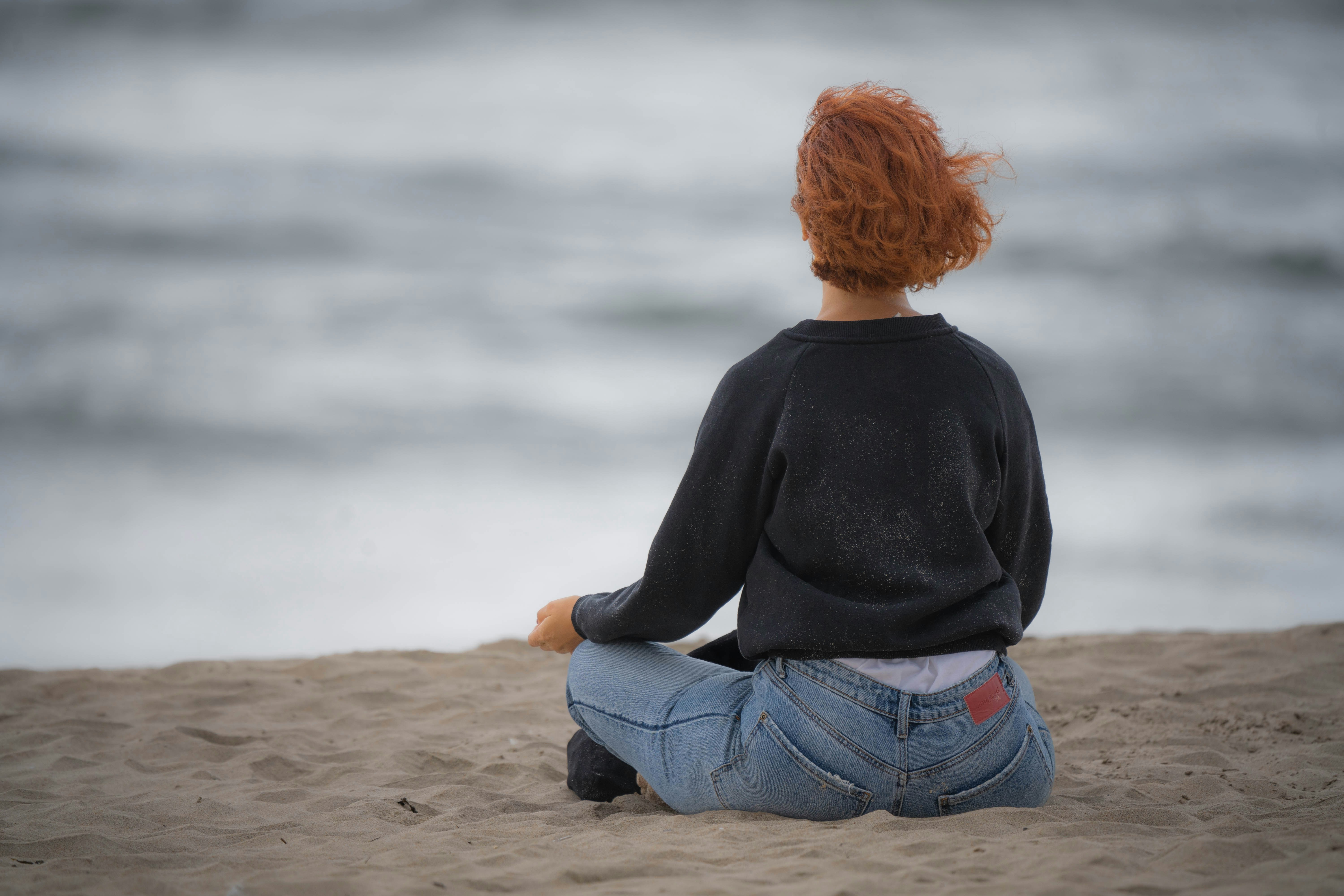 A woman with red hair meditating on the beach, facing the sea with her back to us.