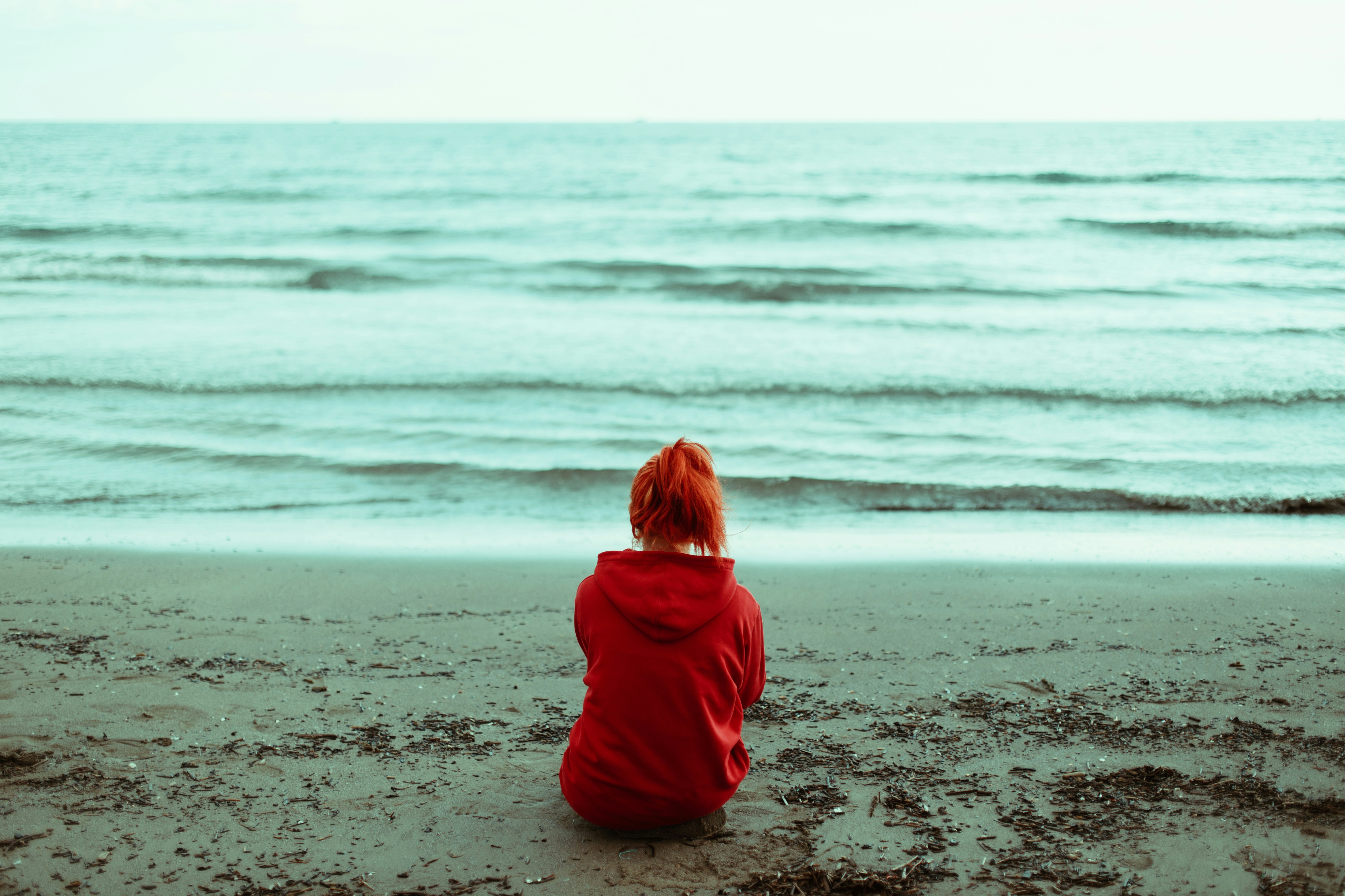 A person with red hair and wearing a red hoodie sits facing the ocean.