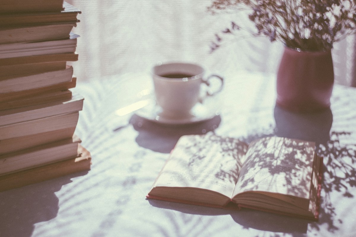 An open book, a stack of books, a mug of tea and a vase of flowers sit on a desk.