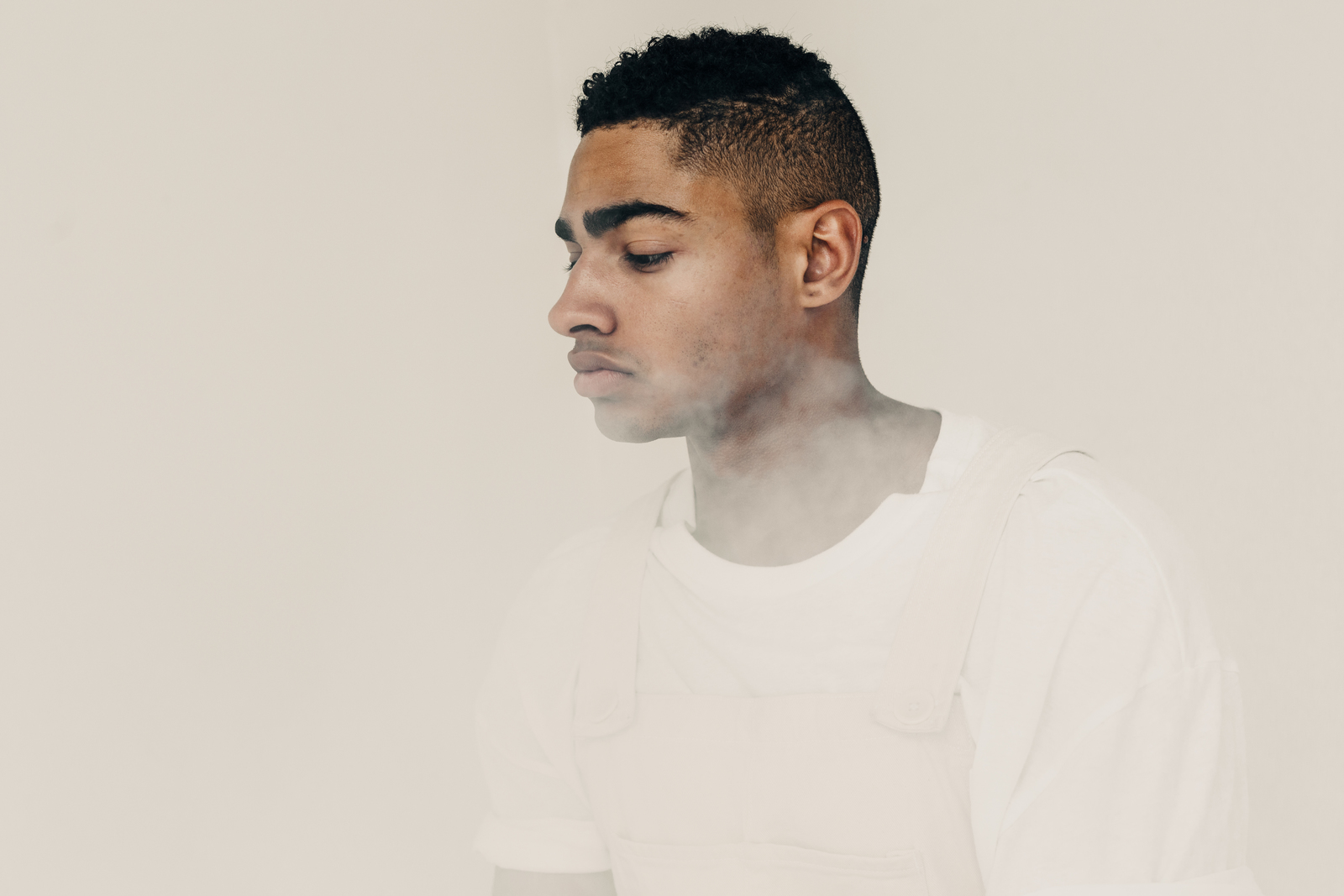 A Black man looking down, wearing a white t-shirt in front of a white background.