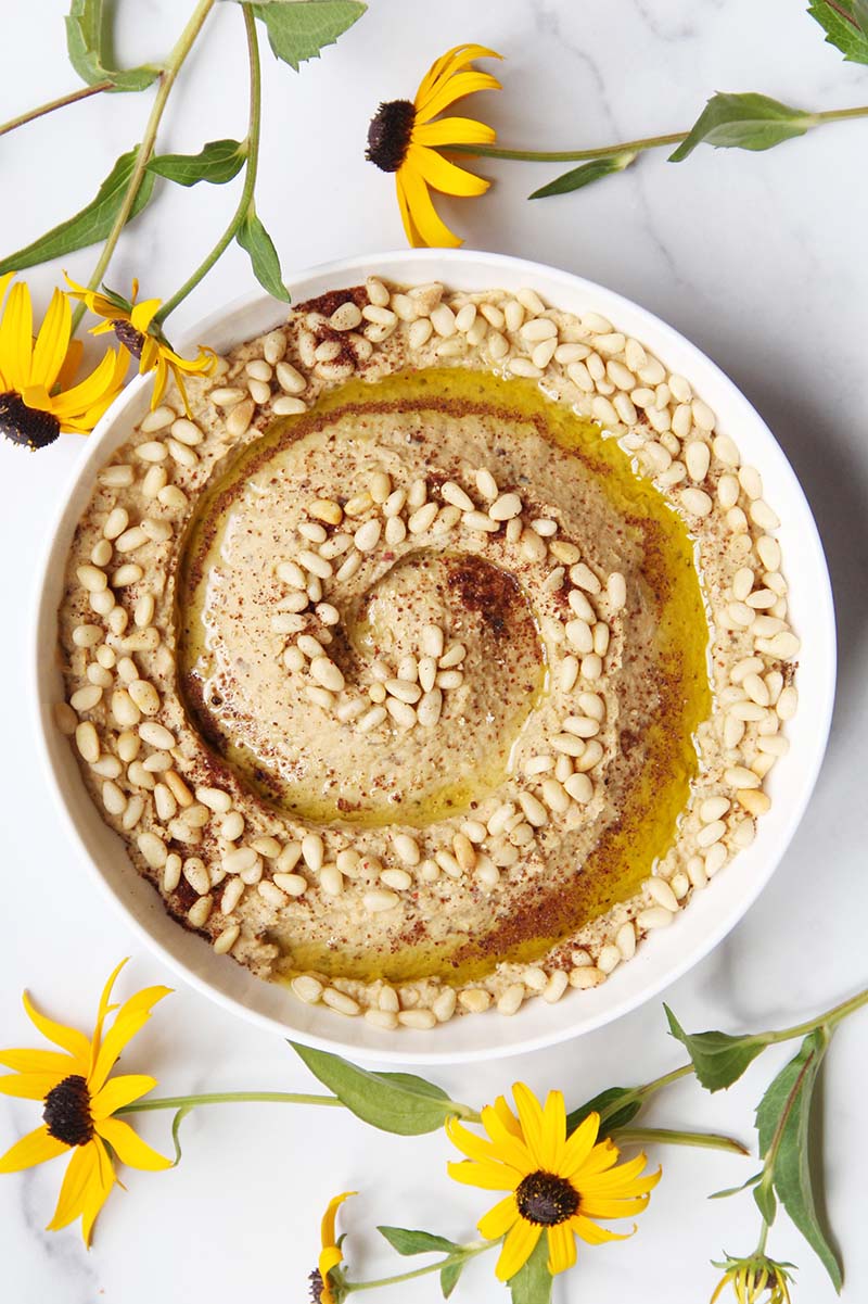 Serving bowl of eggplant hummus, shown on a white background with late-summer flowers.