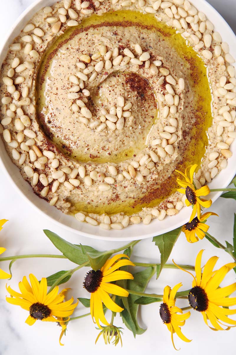 Serving bowl of eggplant hummus garnished with olive oil, pine nuts, and sumac, shown on a white background.