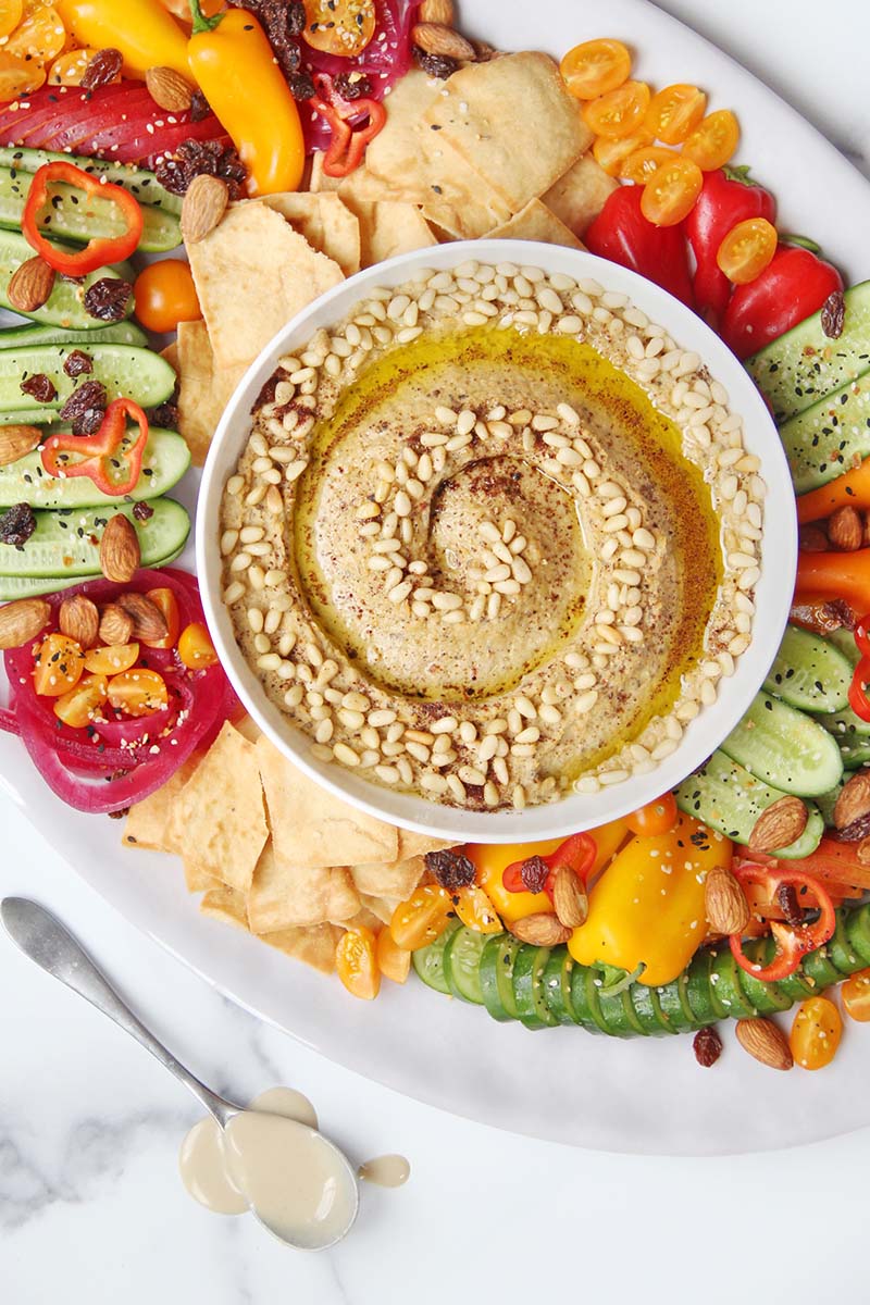 Colorful mezze platter with baba ganoush hummus in the middle, surrounded by pita chips, vegetables, and fruits, shown next to a spoonful of tahini.