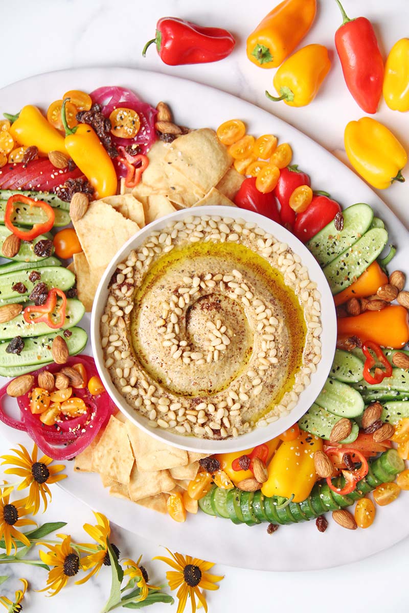 Beautiful eggplant hummus mezze platter, shown on a white background with colorful flowers and vegetables.
