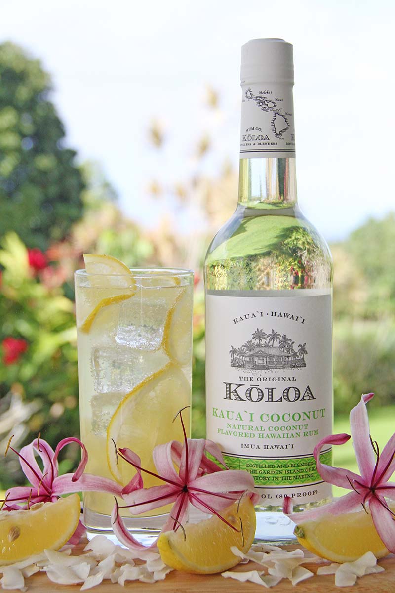 Glass of fizzy coconut rum lemonade, shown next to a bottle of coconut rum in a tropical garden.