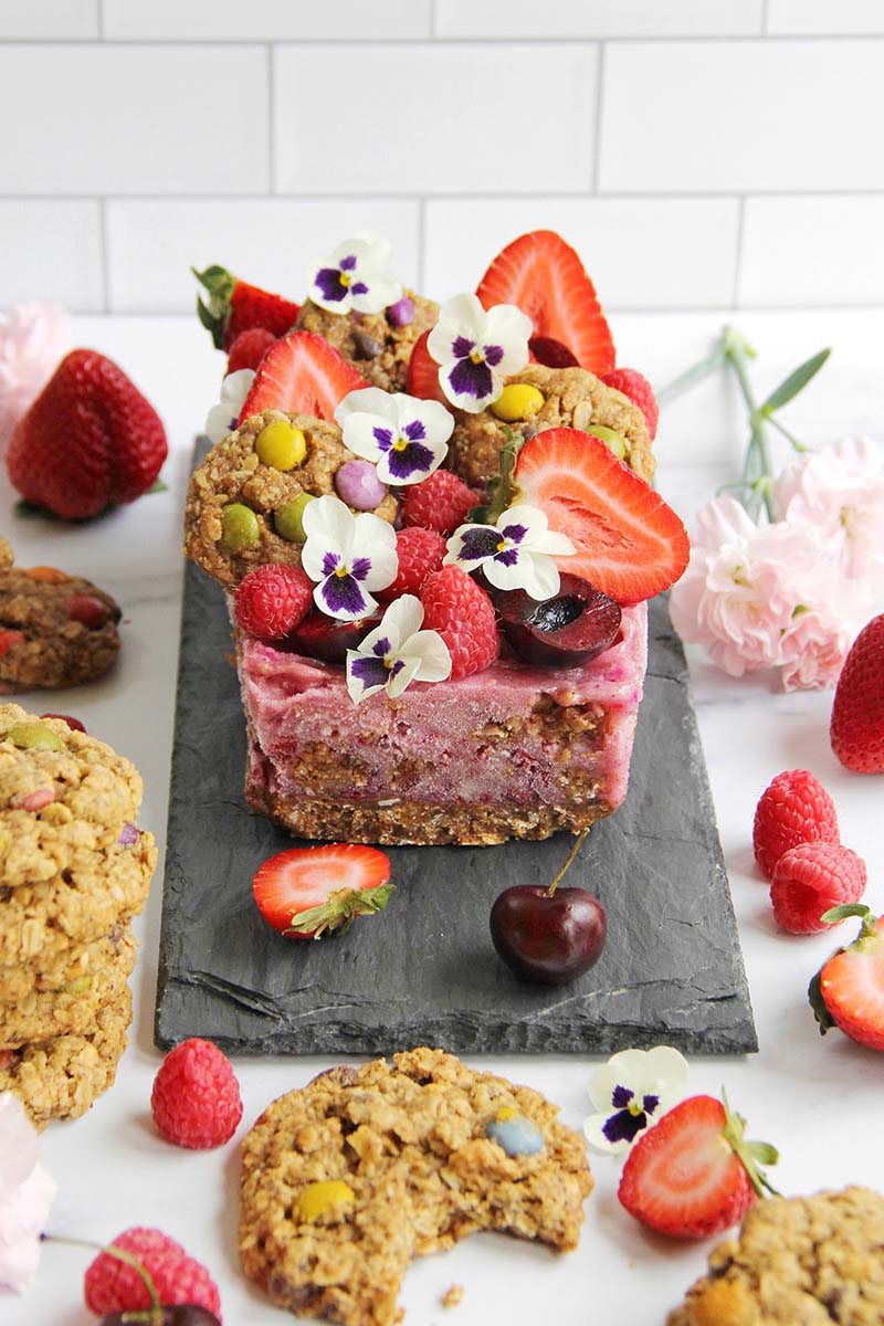 Beautiful and healthy berry nicecream cake topped with fresh summer berries, edible flowers, and miniature cookies.