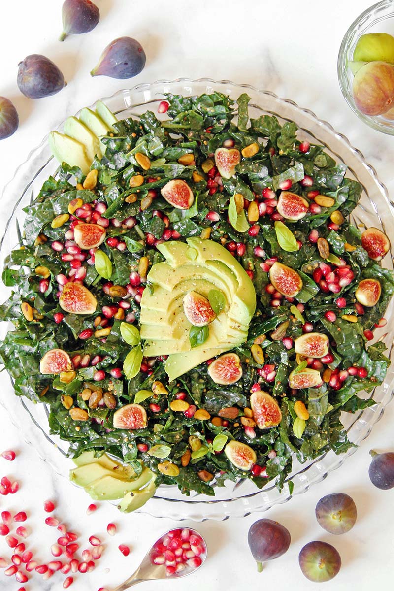 Beautiful fig, avocado, and pomegranate kale salad topped with pistachios and basil, shown with fresh figs.