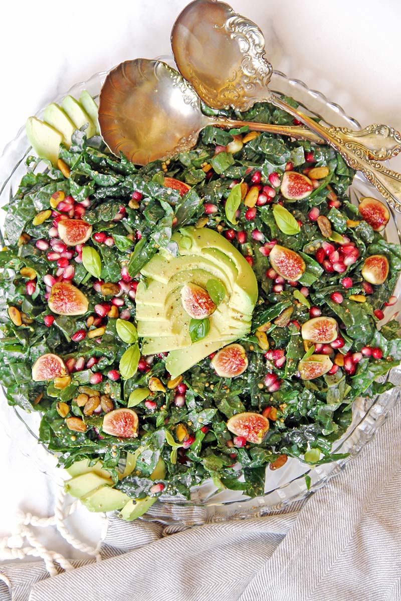 Fig, avocado, and pomegranate kale salad on a serving dish, shown with vintage silver serving spoons on a white background.